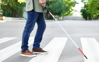 Close up of someone's legs walking across a crosswalk, with a white cane held out in front of them.