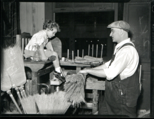 A photo of an early Lighthouse worker making brooms speaking to a young girl visiting the factory