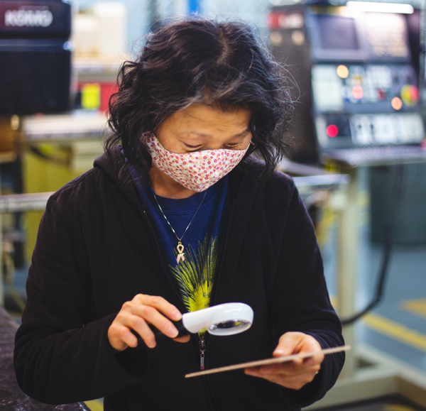 Candid image of an employee with a medium dark skintone and black hair using a handheld magnifying glass to look at a card she is holding in her hand. She is wearing a face mask.