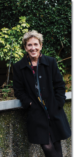 A medium wide portrait of Jane Elliot, a light skinned woman with short light hair. She is standing in a garden, leaning against a rock wall, smiling.