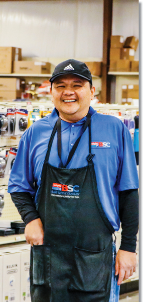A medium portrait of Mike Soriano, a man with a medium dark skintone. He is smiling and standing inside of a BSC store with office products behind him, and wearing a BSC shirt and apron.