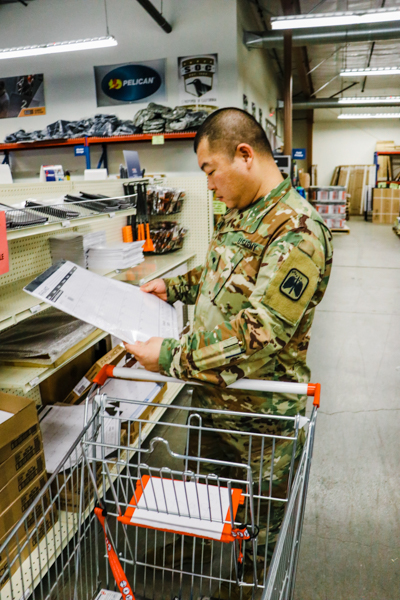 A candid image of a military member shopping in a BSC store. The man has medium dark skin and is wearing camouflage fatigues. He has a shopping cart next to him and is holding a large desk calendar in his hands.