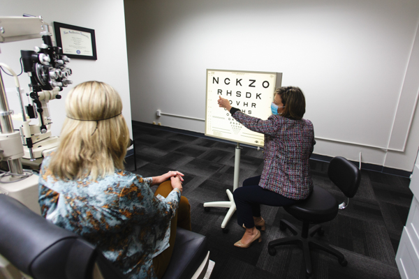 Image of an eye exam in process. One woman with her back to the camera is sitting in an exam chair. Dr. Shagas is sitting next to a large eye chart, pointing to a row of letters.