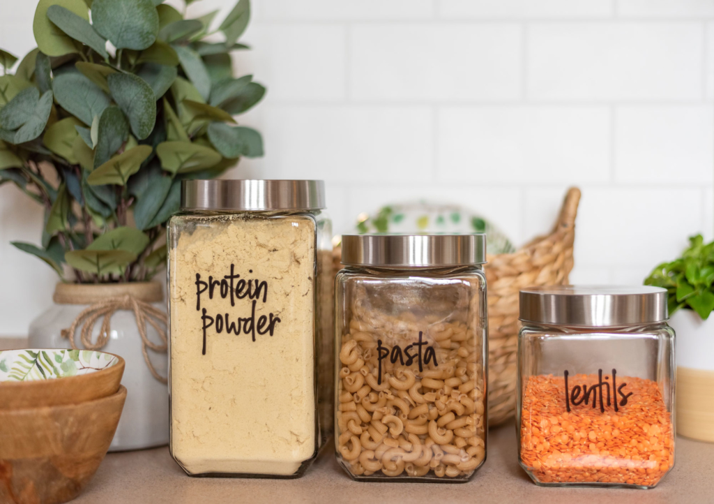 Jars on top of a counter, one with protein powder, second pasta, third lentils