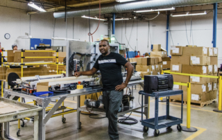 John Koigi stands in a machine shop next to a machine.