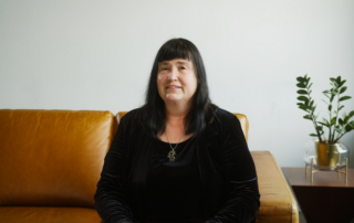 Portrait of Carla, a light-skinned middle aged woman smiling and sitting on a couch