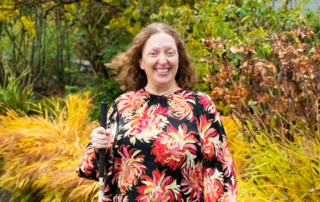 Portrait of Melinda, a light-skinned middle aged woman wearing a floral top and holding a white cane in a garden and smiling