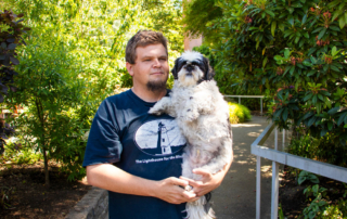 Portrait of Nick, a light-skinned man in his 30s holding a small dog guide and standing and smiling in a garden in Seattle