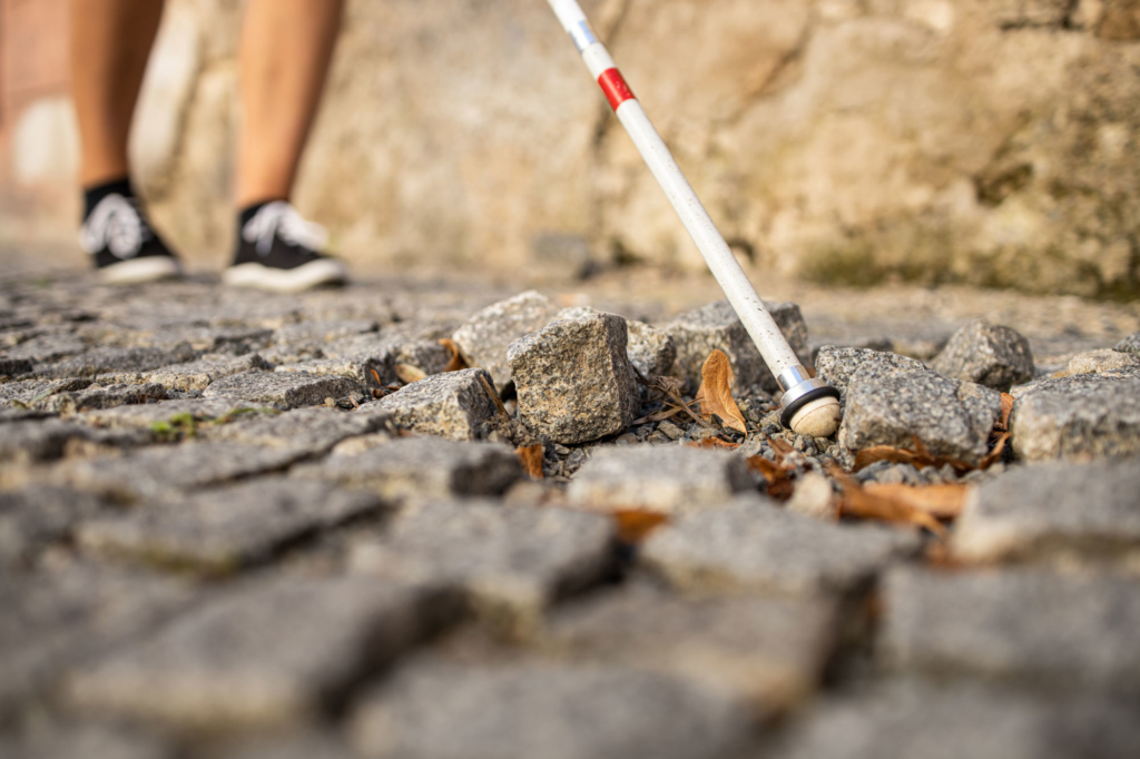 Up close image of a cobblestone sidewalk with the tip of a white cane over an area where the rock is loose.
