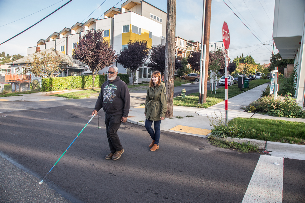 Two people walking across a residential street, one is using a white cane while the other follows closely behind.