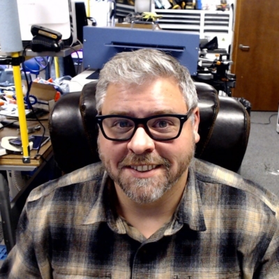 Portrait of Ian, a light skinned man with thick rimmed glasses, salt and pepper hair and beard smiling at the camera.