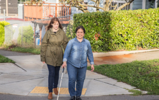 Two women walking down a sidewalk, one is using a white cane.