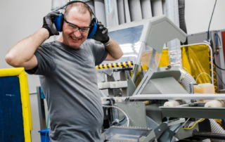 John Cashion, a light skinned man, is wearing large ear protection headphones, clear safety glasses, and is laughing while standing next to a large machine.