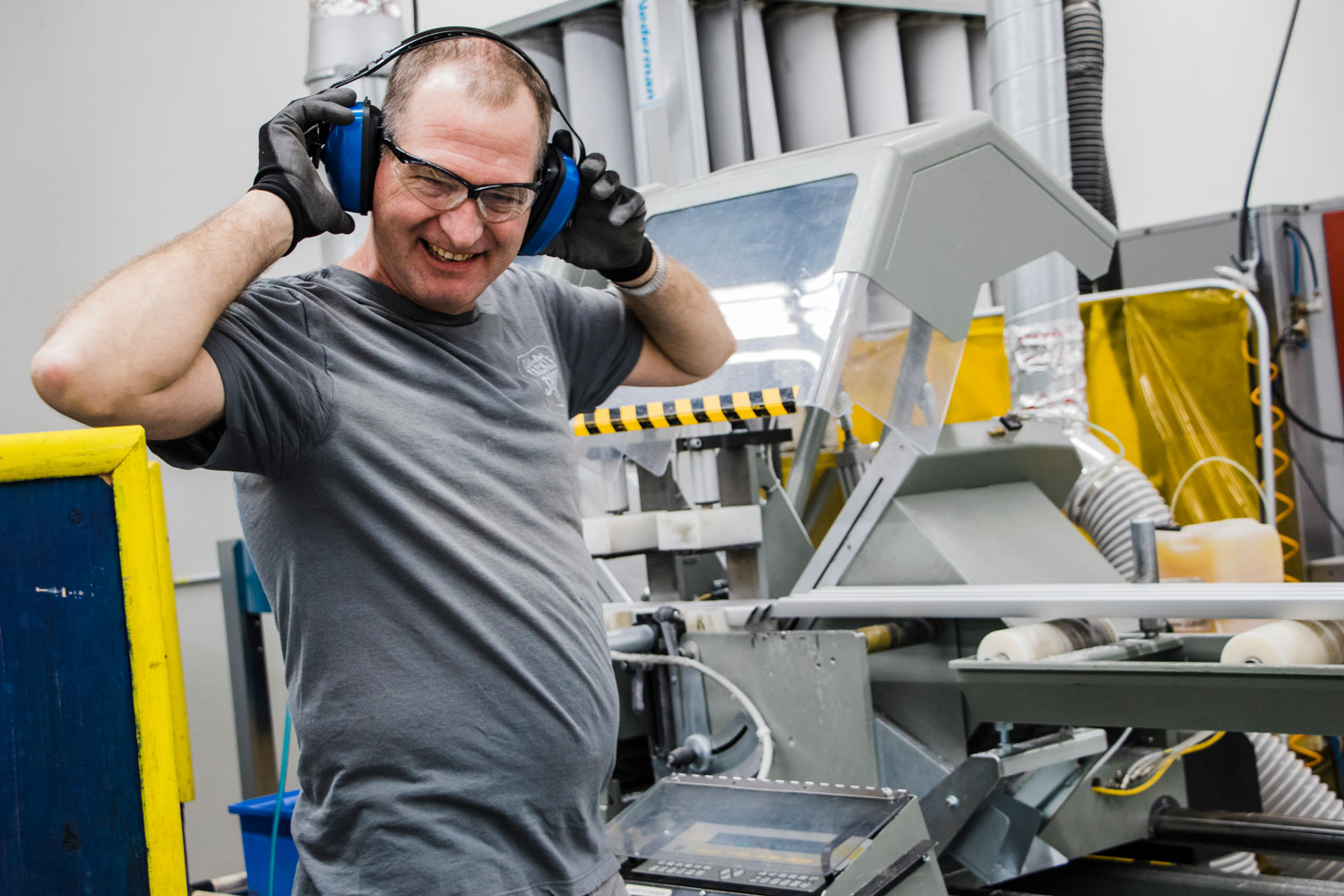 John Cashion, a light skinned man, is wearing large ear protection headphones, clear safety glasses, and is laughing while standing next to a large machine.