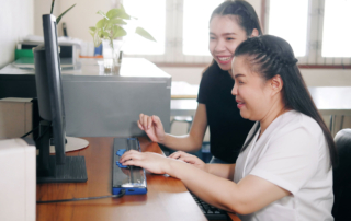 Two women sit in front of a computer. One has both her hands on a refreshable braille display.