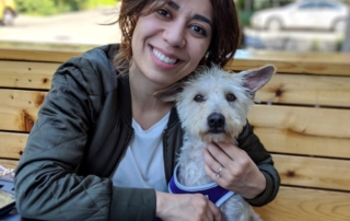 Roya, a dark haired woman smiling with her small white dog.