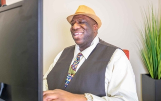 A dark skinned man wearing a hat, vest, and tie and sitting in front of a computer, smiling.