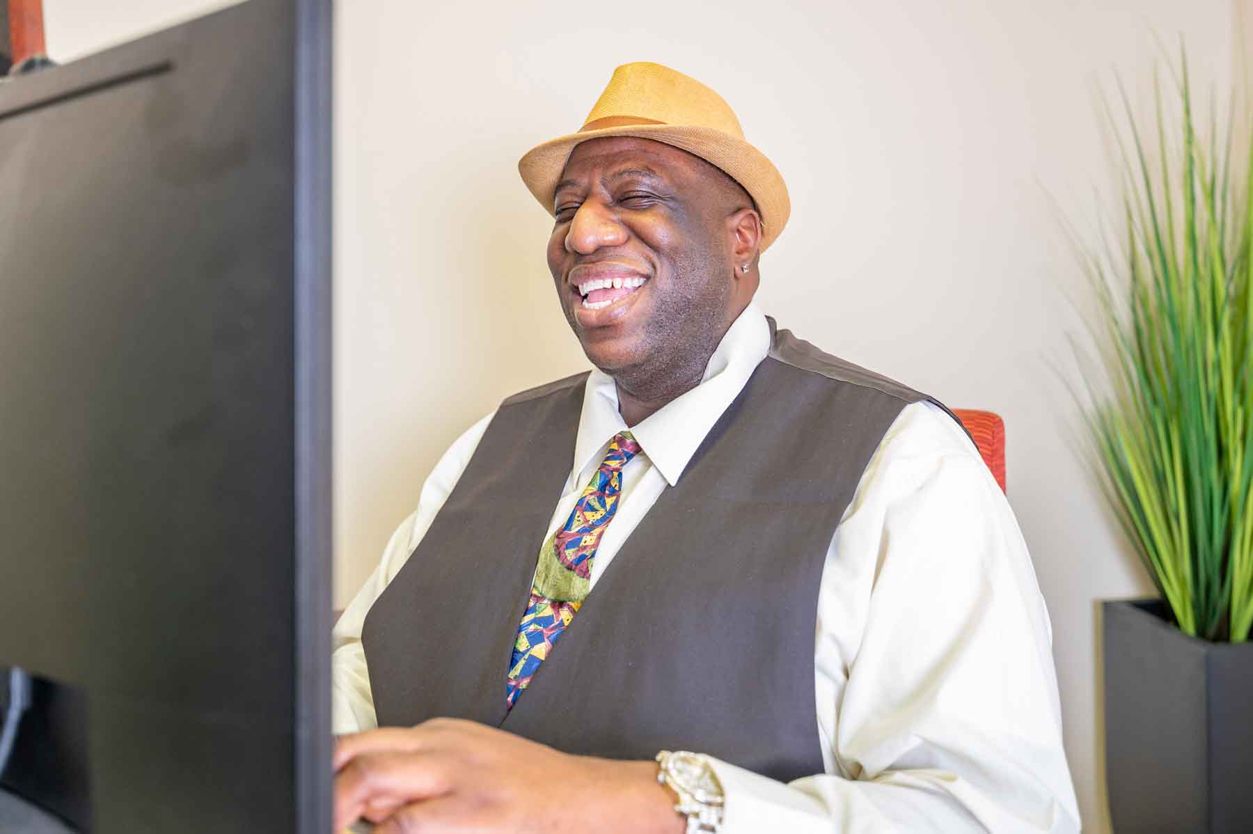 A dark skinned man wearing a hat, vest, and tie and sitting in front of a computer, smiling.