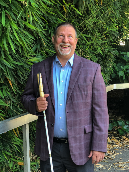 George Abbott smiling in a garden holding a white cane