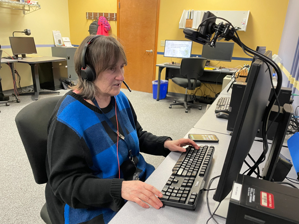 Gaylen Floy, Computer and Assistive Technology Instructor, a light skinned middle aged woman, sitting at a computer and wearing a headset.