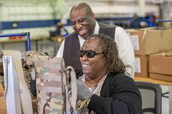 Katine Gadsden, Production Worker, a dark skinned woman with curly hair is wearing sunglasses and sitting at a work table holding a camoflauge water bladder in her hand. Behind her, Earnest Glover, Marketing Assistant is standing and smiling.
