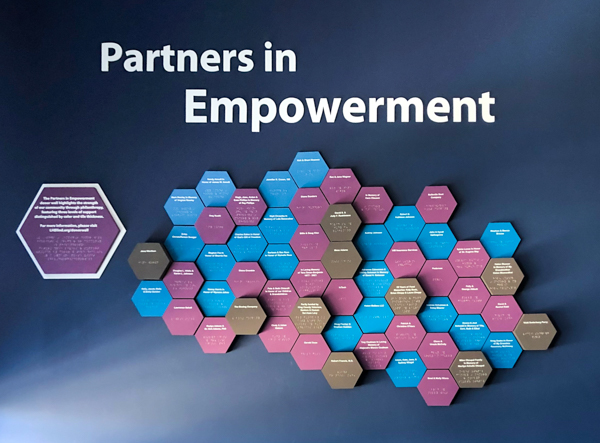 Partners in Empowerment Donor Wall. A dark blue wall with grey, light blue, and purple honeycomb shaped tiles across it.