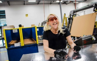 A woman with light skin and blonde hair is smiling joyfully. She is in a warehouse and is wearing dark eye protection as well as gloves.