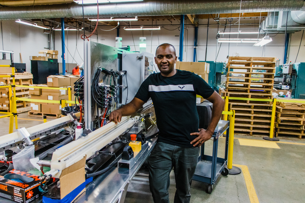 A dark skinned man standing proudly in front a machining workstation in Seattle