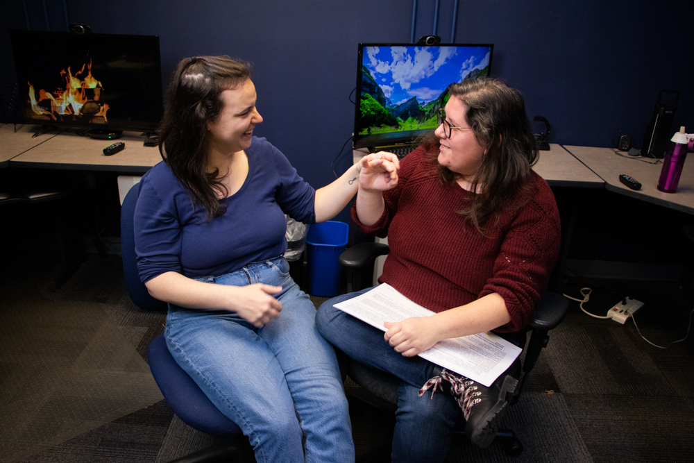 A dark-haired DeafBlind woman uses tactile ASL to communicate with a female interpreter in a Lighthouse computer lab in Seattle