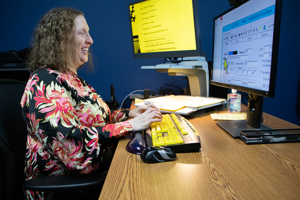 A light-skinned woman sits and types at a computer workstation outfitted with a CCTV and a large print yellow keyboard