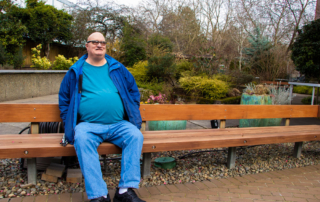 Greg Brown sits outside on a bench in a garden. He is light skinned and bald headed, wearing large glasses.