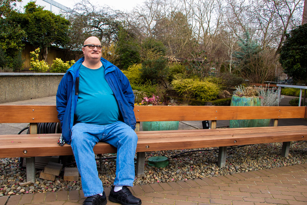 Greg Brown sits outside on a bench in a garden. He is light skinned and bald headed, wearing large glasses.