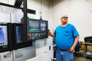 Greg Brown stands next to the control panel of a large machine. He is light skinned, with a bald head, wearing large thick framed glasses.