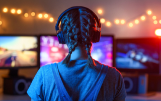 The back of a person's head, wearing large headphones and standing in front of a desktop monitor.