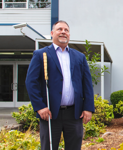 Portrait of George Abbott, a light skinned man with salt and pepper beard and hair. He is standing outside, smiling, and holding a white cane.