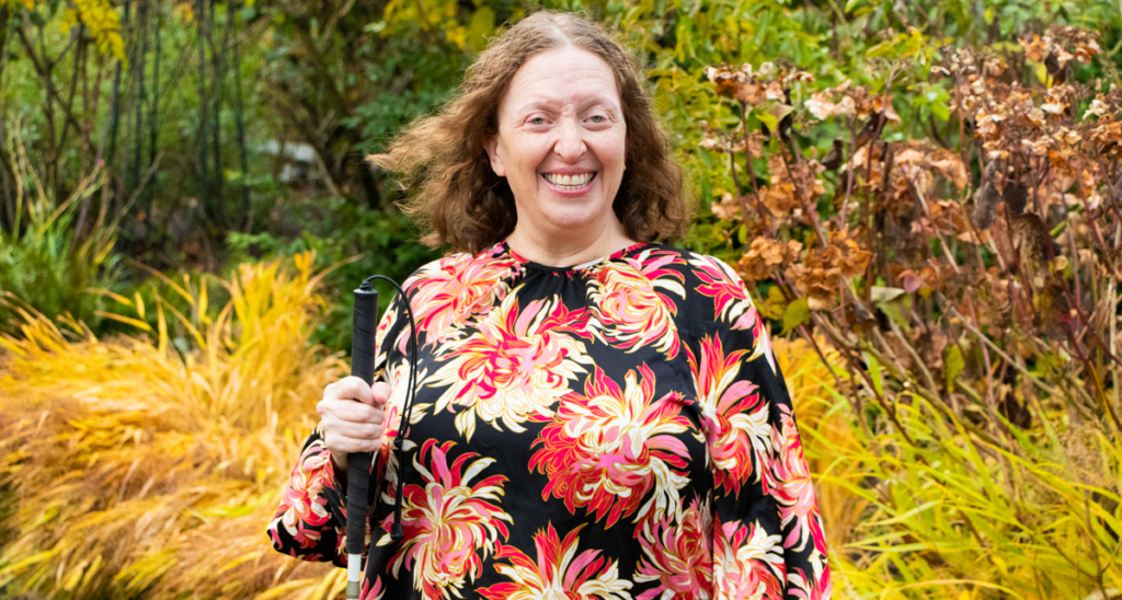 10 – Melinda Slovernick – FY22AR-1 Portrait of Melinda Slovernick, AbilityOne Base Supply Center Administrative Assistant. She has light skin and light hair and it standing in a garden, holding a white cane.