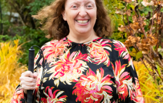 Portrait of Melinda Slovernick, AbilityOne Base Supply Center Administrative Assistant. She has light skin and light hair and it standing in a garden, holding a white cane.