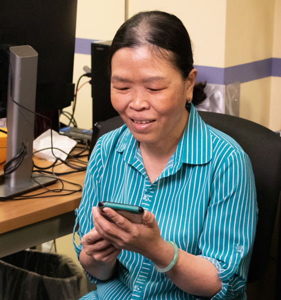 Mai Ho, Production Worker, participating in Computer Training Program classes. She is a medium skinned woman with dark hair pulled back. She's holding a smartphone in her hand and looking at it while smiling.