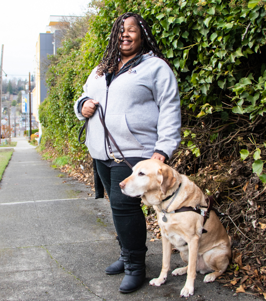 A portrait of Nicole Corbett, a dark skinned woman with long dark braids, standing with her dog guide.