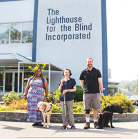 FY2022 Annual Report cover image - pictured - Three people standing outside in front a large blue building with a logo reading, "The Lighthouse for the Blind Incorporated." One woman has dark skin and long dark braids and has a dog guide. The woman in the middle has medium skin, short dark hair, and is holding a white cane. The man to the left has light skin, short dark hair, and has a dog guide.