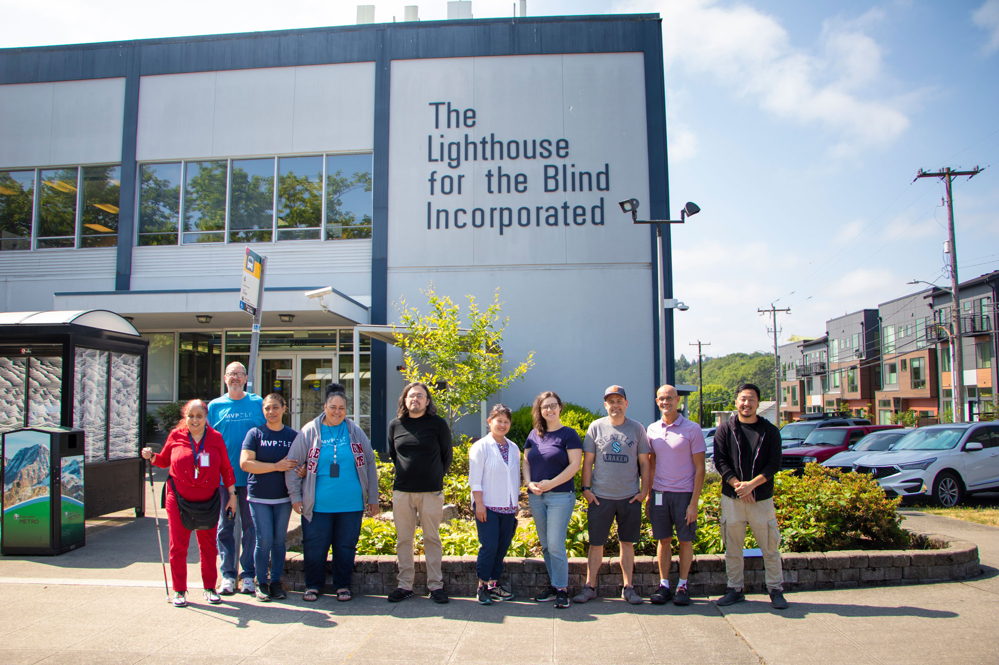 10 people stand outside in front of a large blue building with a logo reading "The Lighthouse for the Blind, Incorporated"
