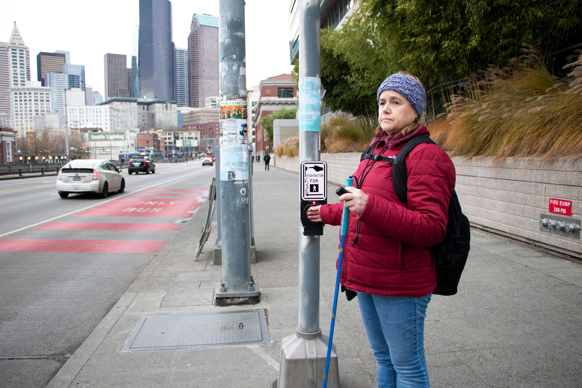 A woman stands at a street corner, with her hand on the accessible pedestrian signal, holding a white cane and a mini guide in her other hand.