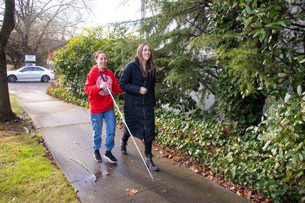 Two young women walking on a sidewalk. One is using a white cane.