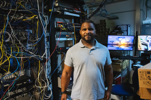 Portrait of Antonio Rozier, a dark skinned man with dark hair. He is standing in a room with wires and computer servers behind him.
