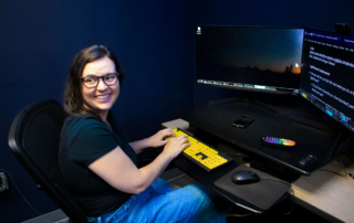 A woman sits in front of a computer, smiling. The keyboard she has her hands on is large print and high contrast, as well as her computer monitor.