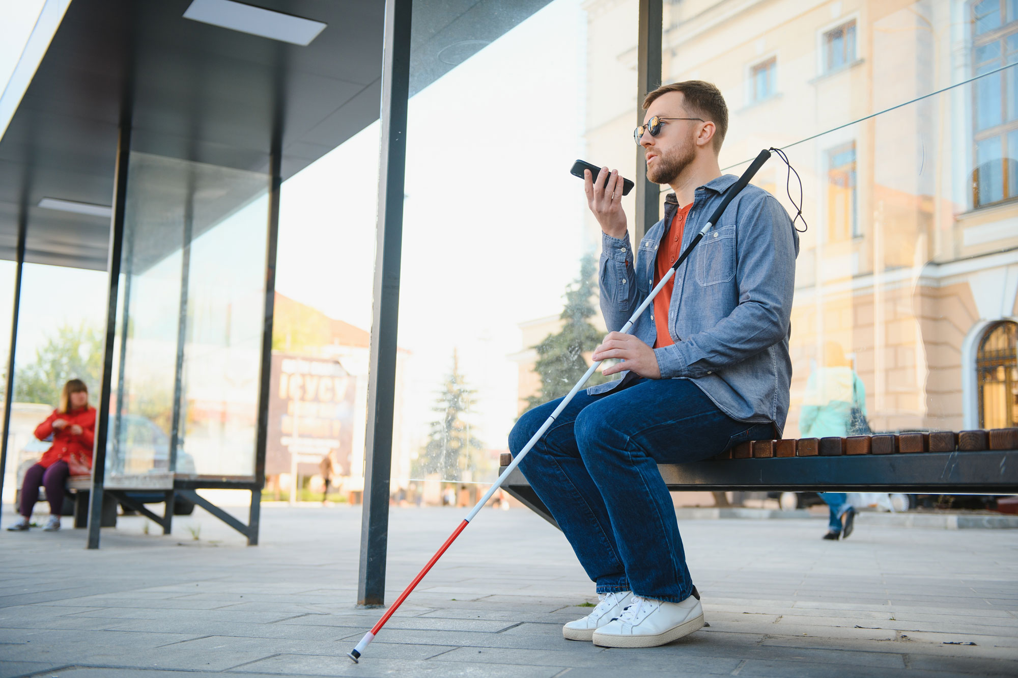 A man sits at a bust stop bench, holding a white cane and using his phone.