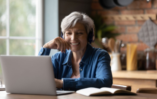 A middle aged woman with grey hair is wearing headphones and smiling at the laptop screen in front of her.