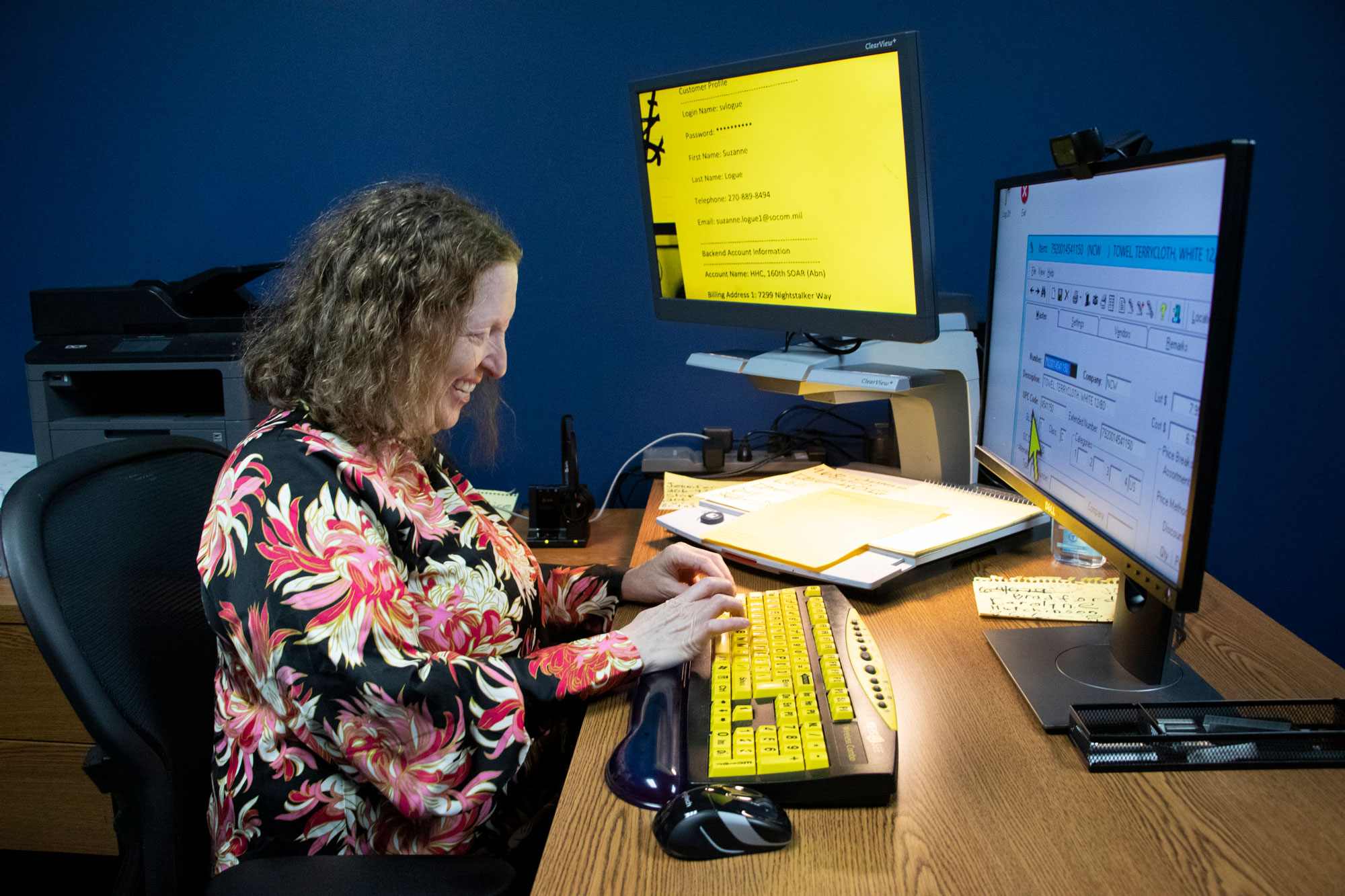 woman sitting at a workstation is typing on a large print keyboard, with two monitors facing her, reflecting magnified content on both