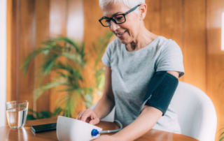 A woman with white hair and dark rimmed glasses sits at a table with a home blood pressure monitor cuff connected to her arm.