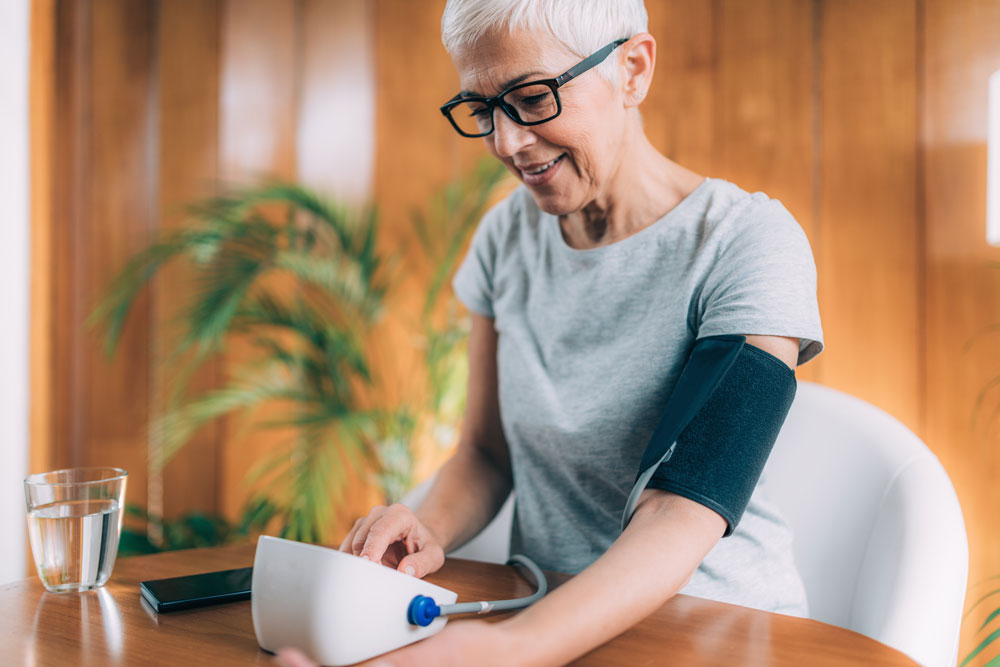A woman with white hair and dark rimmed glasses sits at a table with a home blood pressure monitor cuff connected to her arm.